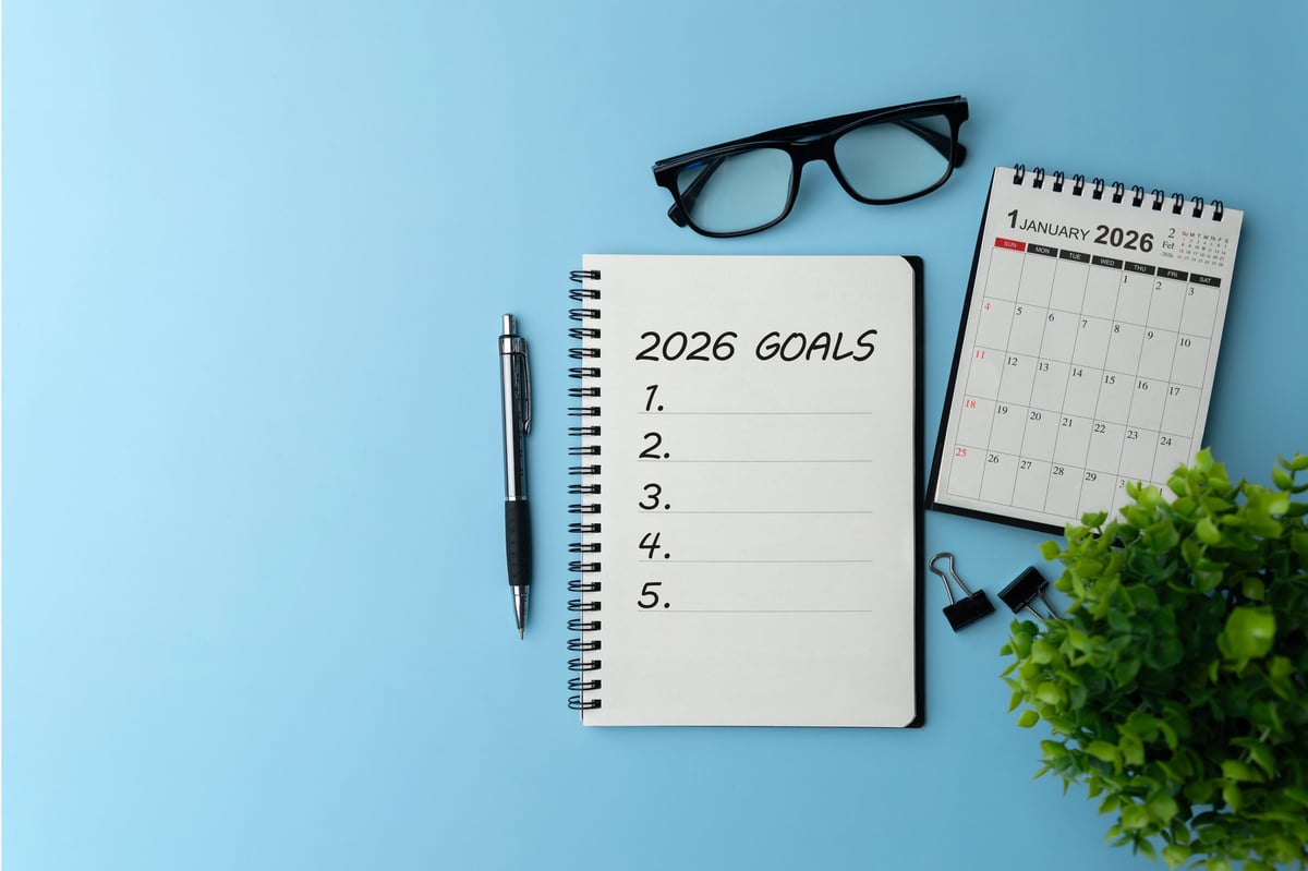 Notebook titled 2026 Goals with a blank checklist on a blue desk beside pen, glasses, mini calendar, representing life and business goal planning, strategy, productivity and a fresh start