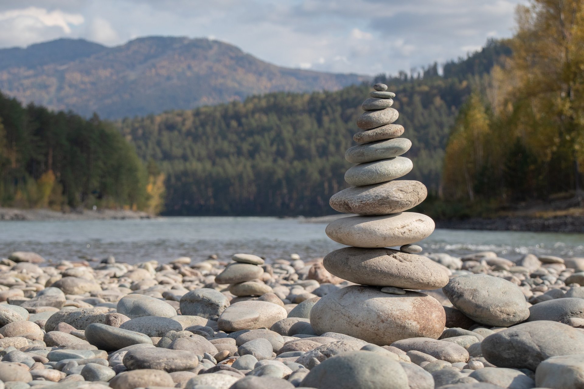 A balanced stone cairn stands on a riverbank, with a backdrop of mountains and trees under a partly cloudy sky.