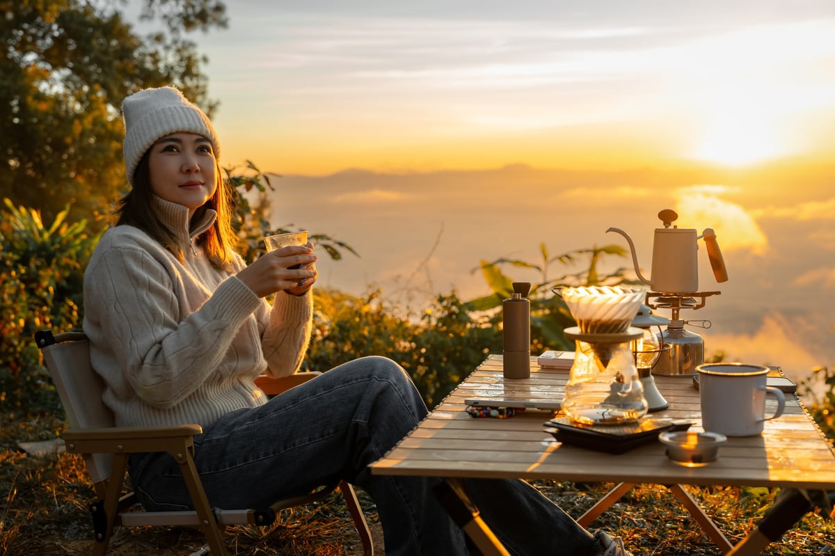 Asian woman drinking hot coffee at campsite in mountains during sunrise. Female traveler solo travel with hiking and camping, sitting at outdoor table with camping equipment, relaxing in nature slow living outdoor lifestyle in forest landscape